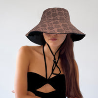 An individual wearing a reversible sun hat with the brown side facing out, featuring a flowery pattern. The black side is not visible, but a black toggle is shown on the chin strap.