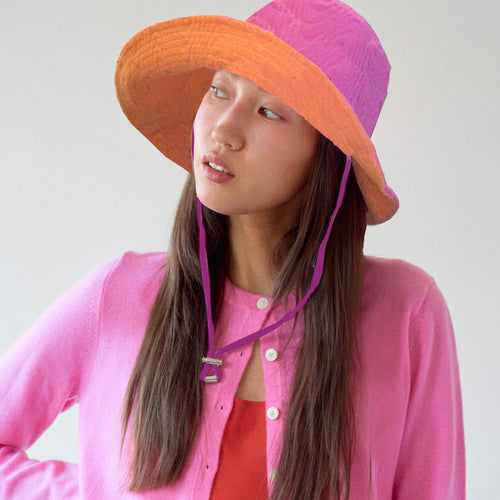 A person wearing a vibrant pink and orange wide-brimmed sun hat with a wavy textured pattern. The hat features an adjustable purple chin strap. She is dressed in a pink cardigan layered over a red top, gazing slightly to the side with a thoughtful expression. The background is neutral and softly lit.