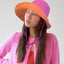 A person wearing a vibrant pink and orange wide-brimmed sun hat with a wavy textured pattern. The hat features an adjustable purple chin strap. She is dressed in a pink cardigan layered over a red top, gazing slightly to the side with a thoughtful expression. The background is neutral and softly lit.