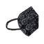 A black and white paisley face mask lying flat on a light plain surface. The mask has a contoured design with a prominent seam down the center and black elastic ear loops.