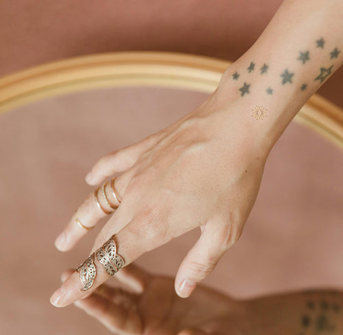 Close-up of a hand of an individual with star tattoos wearing a small adhesive hello sunshine patch with gold sun feature on their wrist.