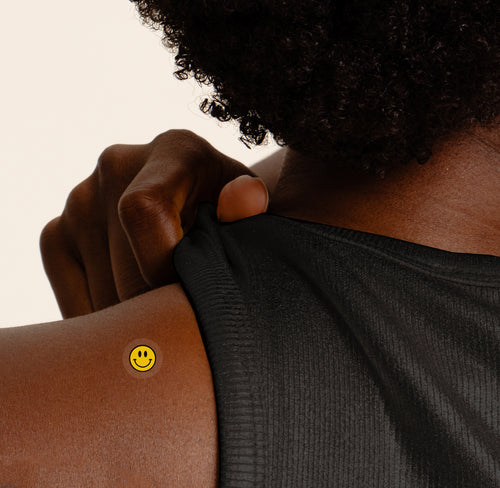 A close-up of a person's shoulder and upper back area with an 'ENERGY BOOST' vitamin patch featuring a small happy face in yellow and black.