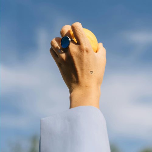  An individual holding up their hand, holding an object with a blue ring on their finger, and wearing a ‘CALL OF BEAUTY’ vitamin patch on their hand.