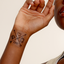A person’s hand with a translucent vitamin patch on the wrist. The design features four symbols arranged in a square pattern: a diamond shape, an outline of a heart, an arrow, and an apple with a small leaf. The symbols are connected by lines forming an ‘X’ shape.