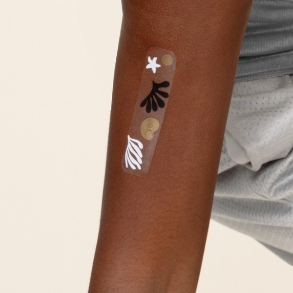 Close-up of an arm with a 'Travel Well'   vitamin patch with black, white and gold shell, star, sea weeds features.