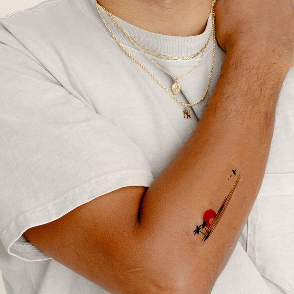 An individual’s folded arm with a vitamin patch. The patch features a small palm tree with two smaller tufts of grass. Above the island, there are three birds in flight. To the right of the palm tree is a sun with seven rays.